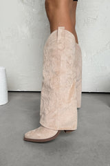 Beige suede cowboy boots worn by a person on a gray floor with a white wall background