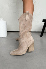 Beige suede cowboy boots on a gray floor with a white vase in the background