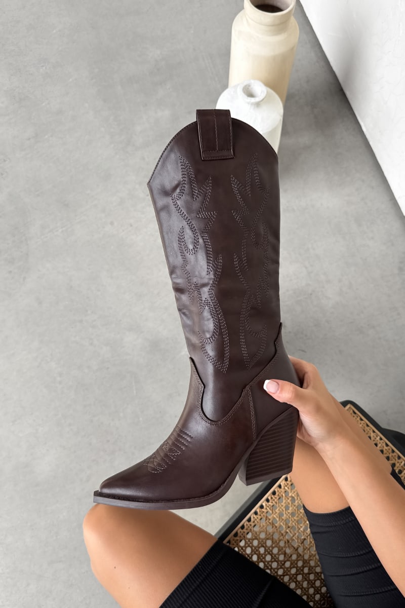 Brown cowboy boot being held on a gray floor with a vase in the background