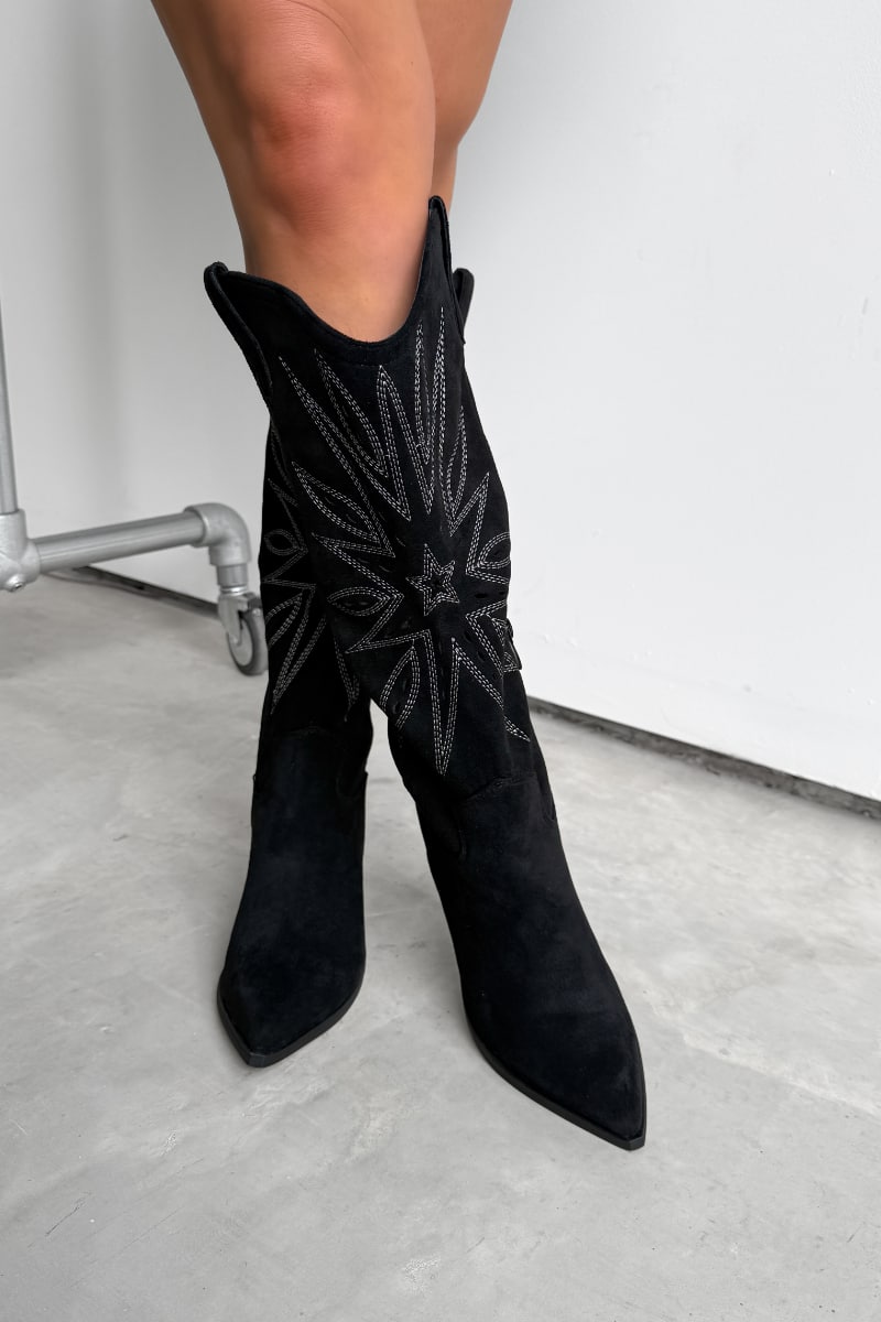 Black knee-high western cowboy boots with a traditional design worn by a person on a light gray floor. 