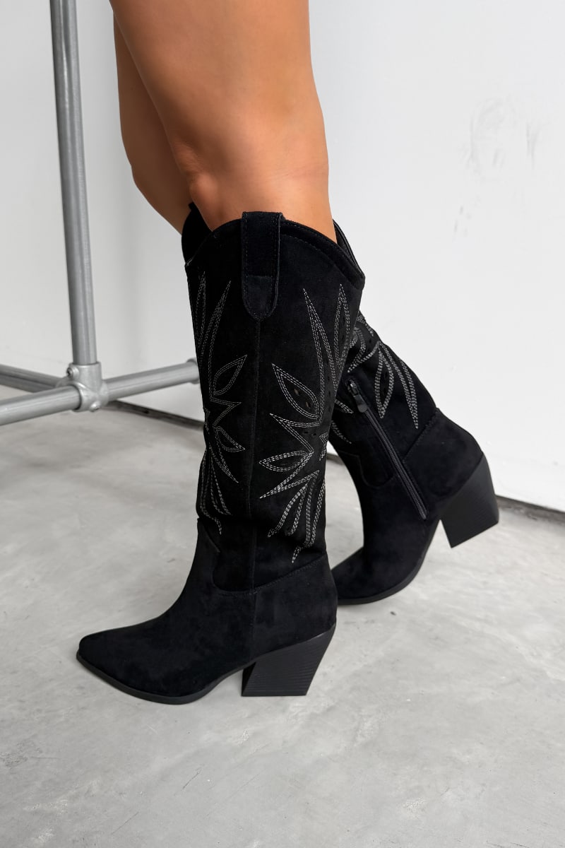 Black knee-high western cowboy boots with a traditional design worn by a person on a light gray floor. - 2
