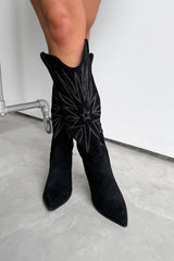 Black knee-high western cowboy boots with a traditional design worn by a person on a light gray floor. 